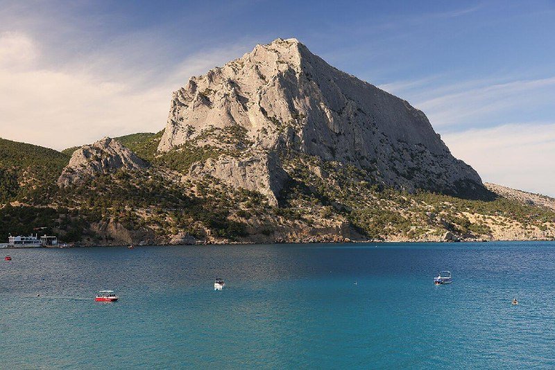 A large rocky mountain rises above a calm, deep blue sea dotted with small anchored boats under a partly cloudy sky.