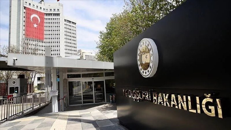 A black sign reading "TC DIŞİŞLERİ BAKANLIĞI" stands beside a building entrance, with a large Turkish flag draped on a nearby high-rise under a partly cloudy sky.