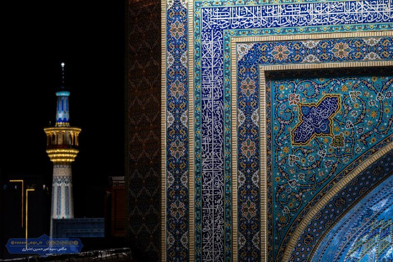 An ornately tiled wall with Arabic calligraphy and floral motifs stands in the foreground at night, with an illuminated minaret visible in the background.