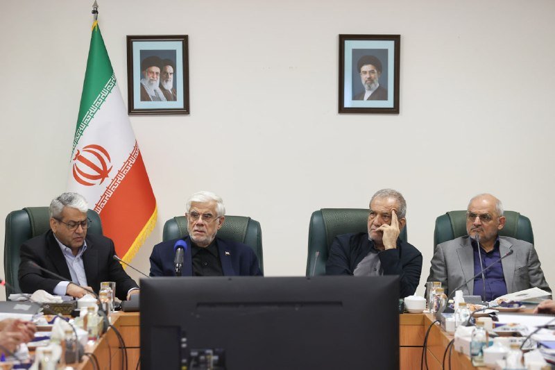 Four men in suits sit at a conference table with microphones, with an Iranian flag and two framed portraits of bearded men in religious attire on the wall behind them.