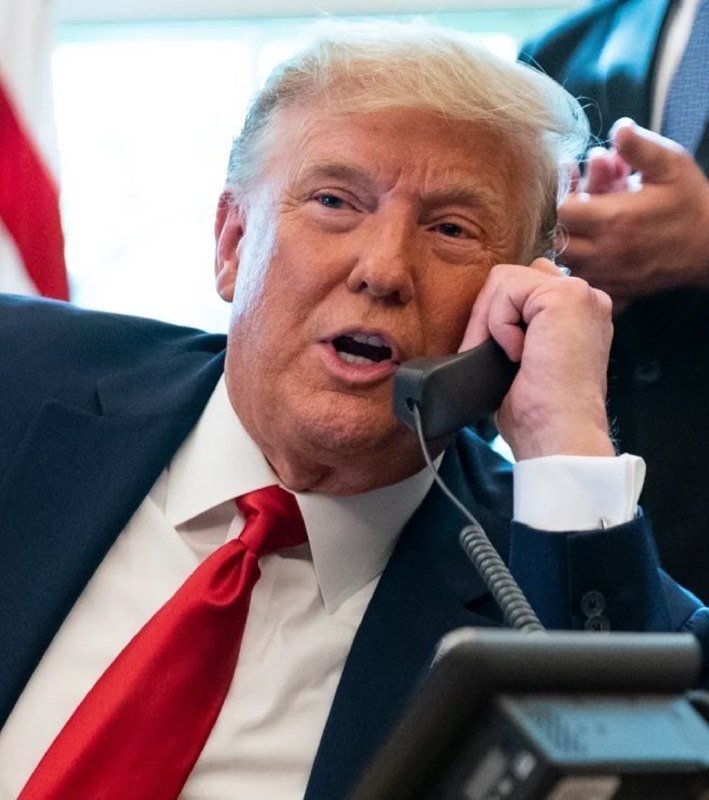 A man with blonde hair in a dark suit and red tie speaks on a telephone receiver, with an American flag visible in the background.