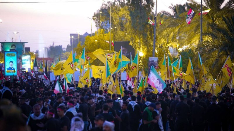 A large crowd marches down a palm-lined street at dusk, waving yellow flags and portraits amid lit billboards.