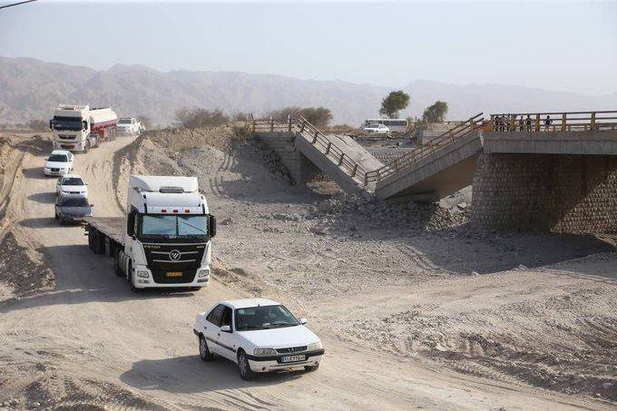 Trucks and cars travel along a dirt road beneath a damaged concrete bridge in an arid, mountainous landscape.