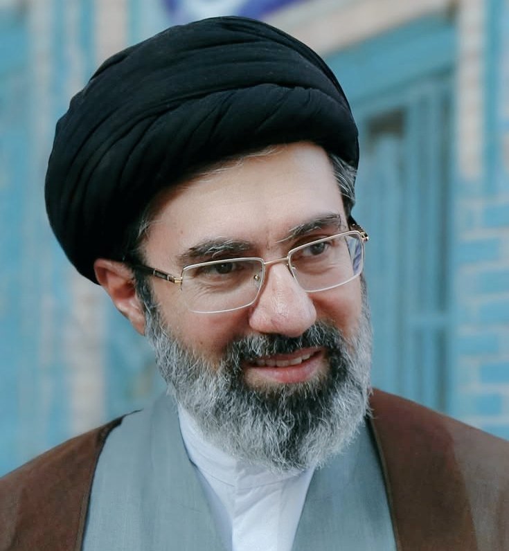 A bearded man wearing a black turban, rimless glasses, and clerical robes smiles in front of a blue-tiled background.
