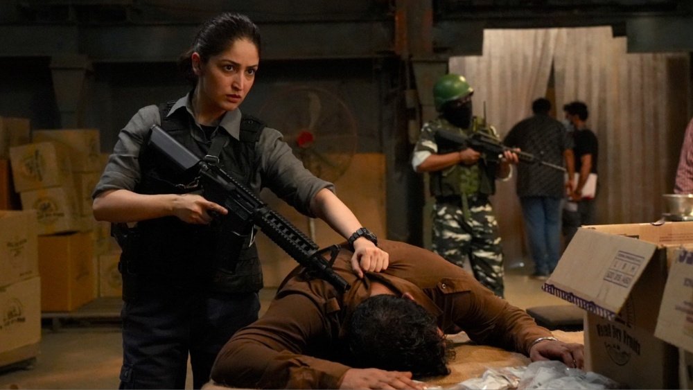 A woman holding a rifle pins a man face-down on a table in a warehouse setting, while an armed soldier in camouflage and other figures stand in the background.