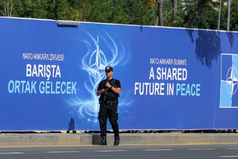 A uniformed officer stands guard in front of a blue banner reading "NATO ANKARA SUMMIT: A SHARED FUTURE IN PEACE."