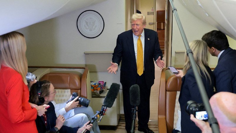 A man in a dark suit and yellow tie gestures with open arms while speaking to journalists holding microphones and cameras inside an aircraft cabin featuring a presidential seal on the wall.