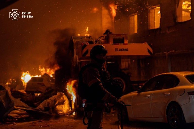A firefighter in protective gear stands amid burning debris and damaged vehicles at night, with a fire truck visible in the smoke-filled background.