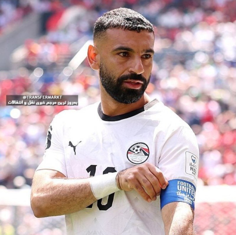 A bearded male soccer player wearing a white Egypt national team jersey with a Puma logo and a captain's blue armband stands on a sunny pitch before a blurred crowd.