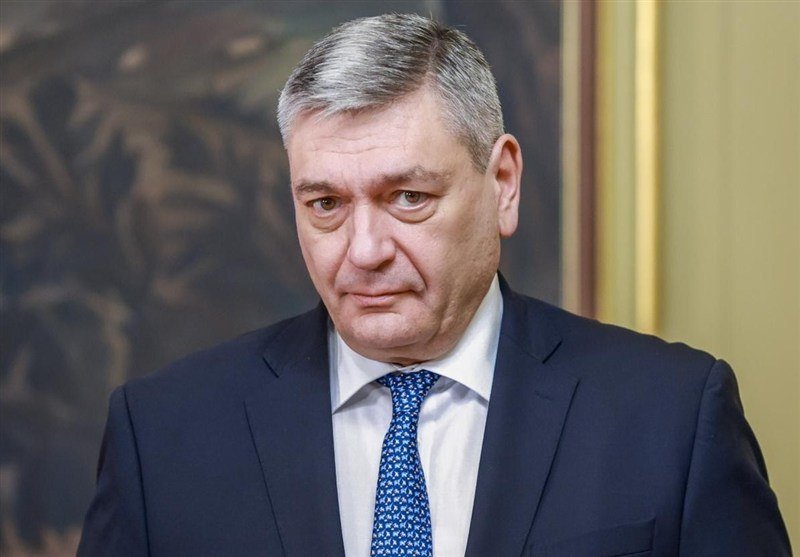 An older man with gray hair wearing a dark suit, white shirt, and patterned blue tie stands indoors in front of a dark painting.