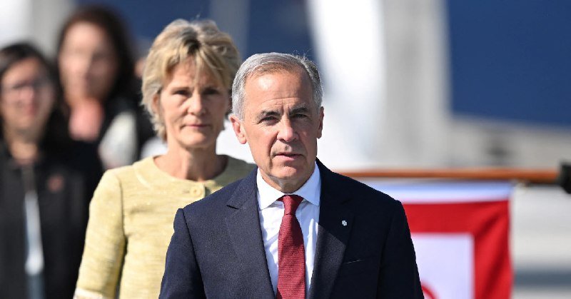 A grey-haired man in a navy suit and red tie stands beside a blonde woman in a yellow jacket, with other people visible behind them near a red and white structure.