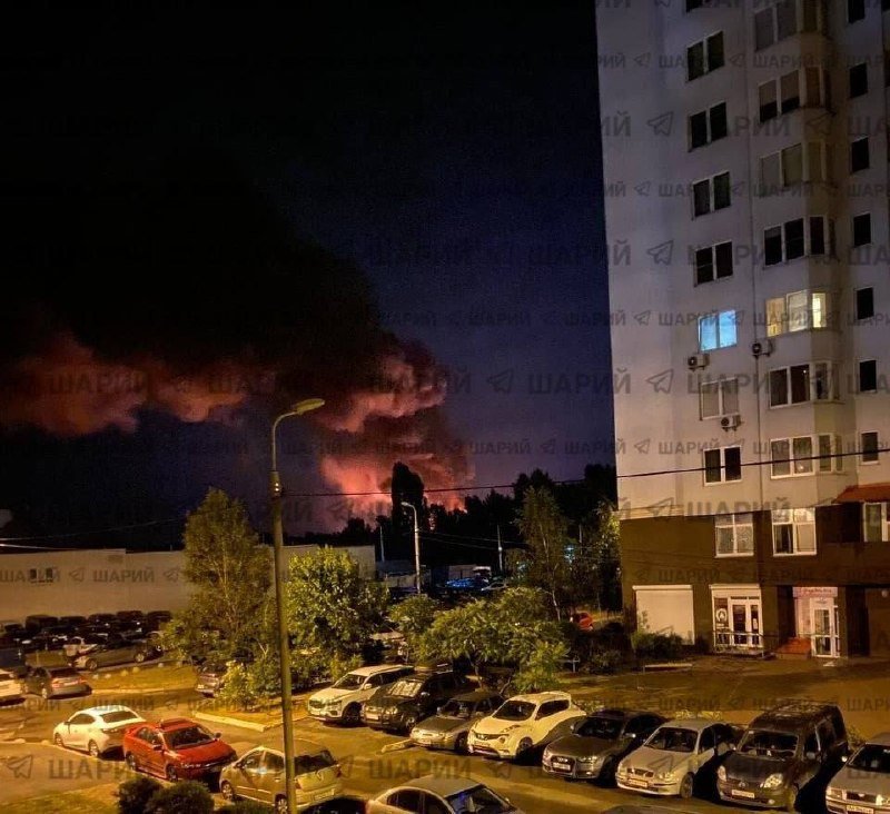 Nighttime photo shows a parking lot filled with cars beside a multi-story apartment building, with a large plume of orange-lit smoke rising over distant trees on the horizon.