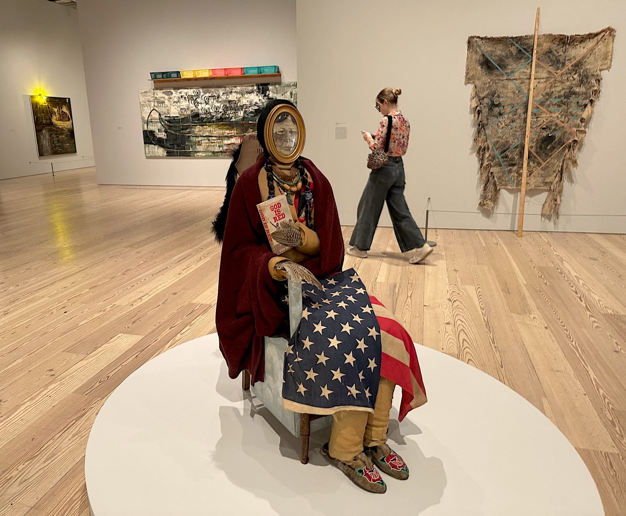 A seated sculpture draped in a red blanket and American flag, holding a "God Is Red" book, is displayed on a circular platform in a gallery.