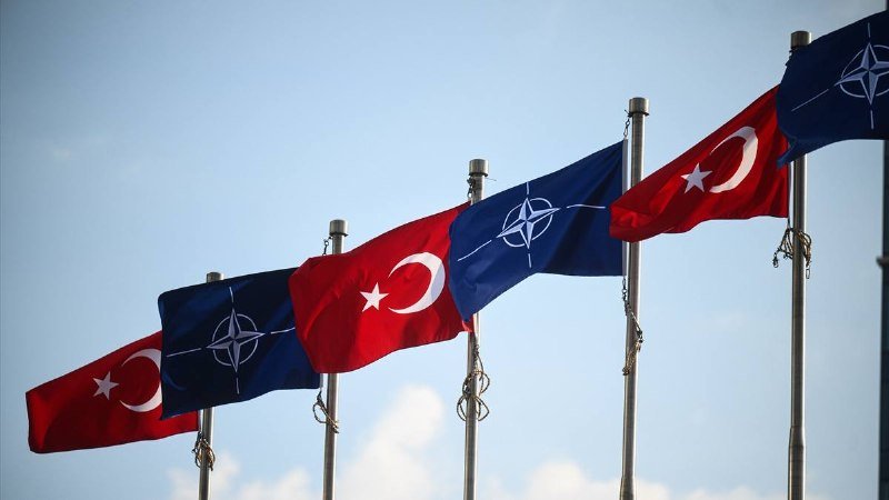 Alternating red Turkish flags and blue NATO flags fly from flagpoles against a clear blue sky.