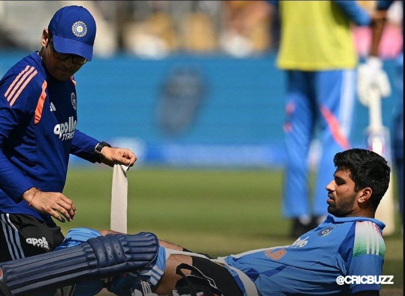 A person in a blue jersey and cap wraps a bandage around the knee of a seated cricketer wearing batting pads, on a cricket field.