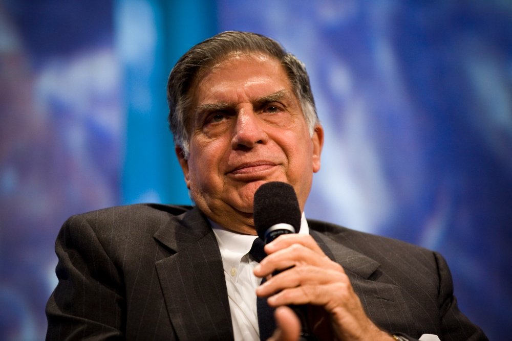 An older man in a dark pinstriped suit holds a microphone and looks upward against a blurred blue background.