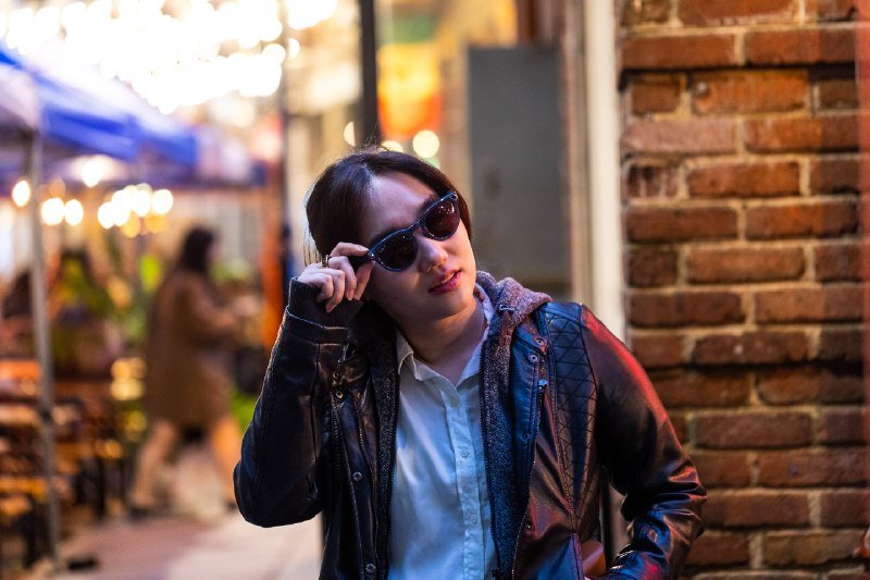 A woman in a black leather jacket and sunglasses poses beside a brick wall at night, adjusting her glasses with one hand.
