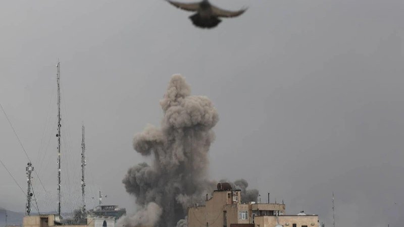 A large plume of gray smoke rises above city rooftops, with a bird flying overhead and communication towers visible on the left.