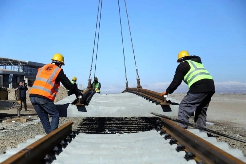 Workers in high-visibility vests and hard hats guide a large concrete railway sleeper suspended by cables onto tracks under construction on an open site.