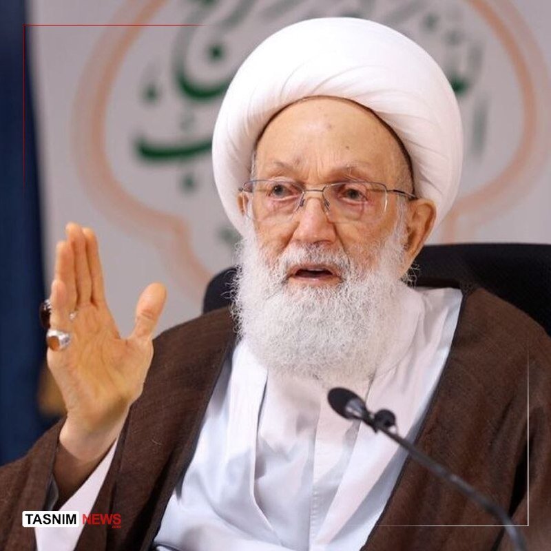 An elderly cleric with a white beard, glasses, and white turban speaks into a microphone, gesturing with his ringed hand, bearing a "TASNNIM NEWS" watermark.