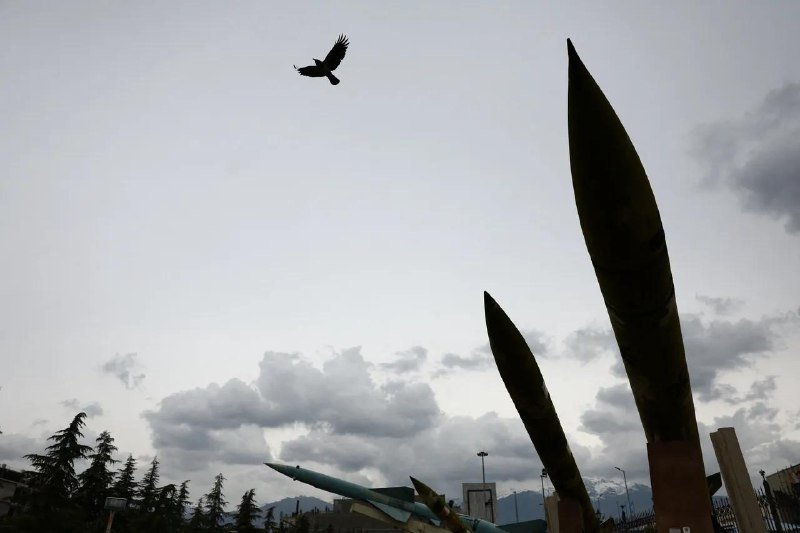 A silhouetted bird flies over two tall missile-shaped monuments displayed outdoors against a cloudy sky.