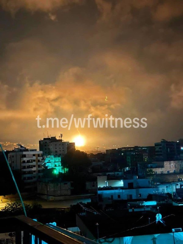 A nighttime cityscape shows illuminated buildings and rooftop water tanks beneath a cloudy sky, with an orange flash and rising smoke visible on the horizon.