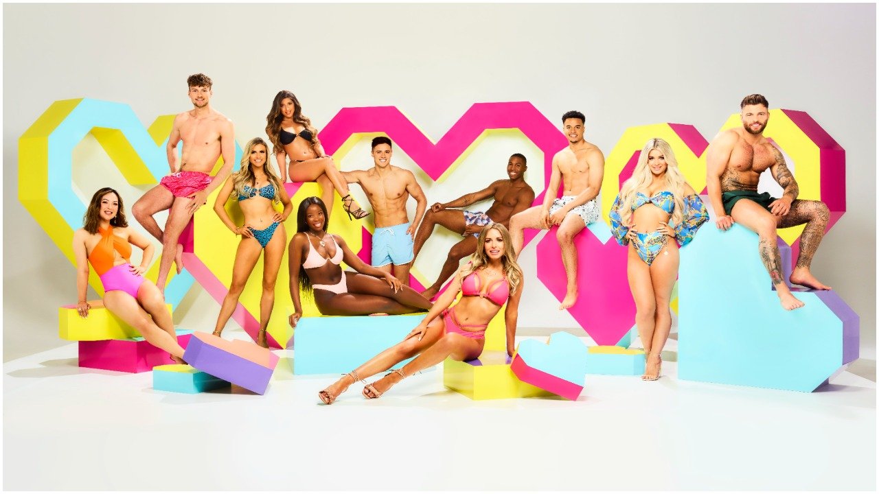 Eleven people in swimwear pose among large, brightly colored geometric blocks forming a heart backdrop in a studio setting.