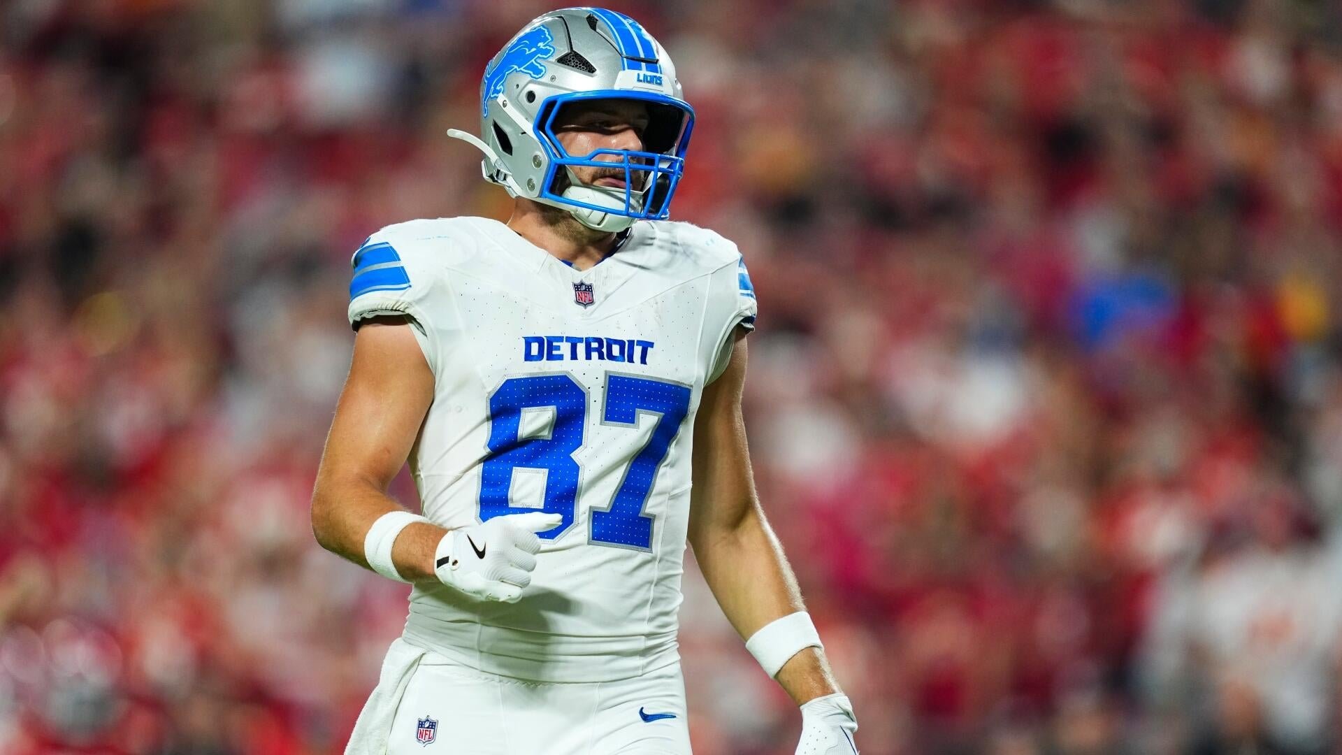 A Detroit Lions football player wearing jersey number 87 walks on the field, set against a blurred crowd of red-clad spectators.