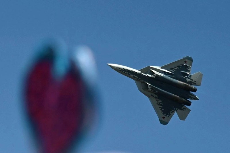 A twin-engine fighter jet with digital camouflage flies through a clear blue sky, while an out-of-focus person in red appears in the left foreground.