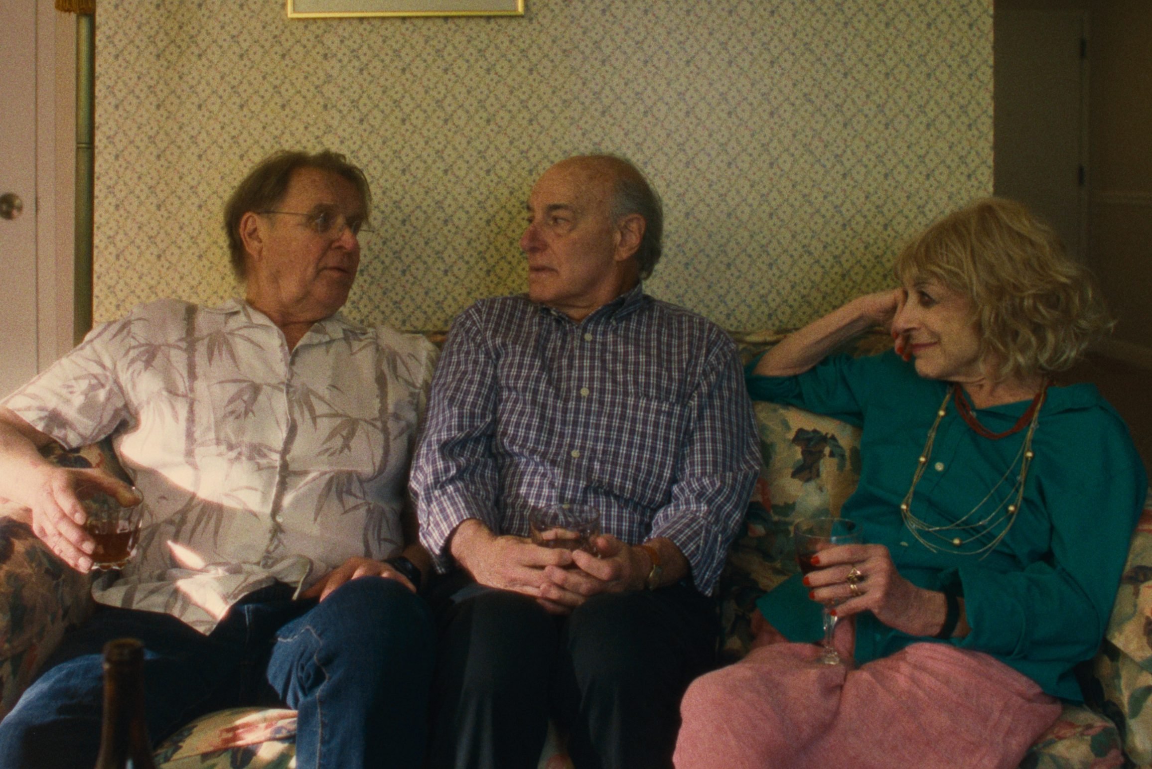 Three people sit on a floral couch holding drinks, engaged in conversation.