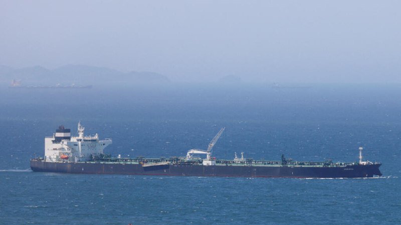 A large oil tanker with a white superstructure and onboard crane sails through open blue waters, with hazy mountains and another distant vessel visible on the horizon.