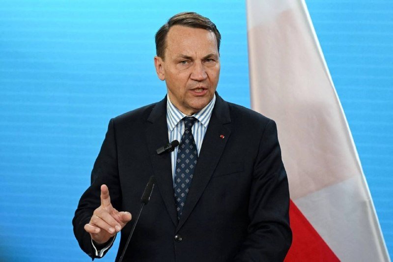A man in a dark suit and patterned tie speaks at a microphone, gesturing with one hand, standing in front of a white and red flag against a blue backdrop.