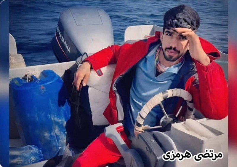 A bearded man in a red jacket and backward cap sits at the helm of a small motorboat on the water, Persian text visible in the corner.