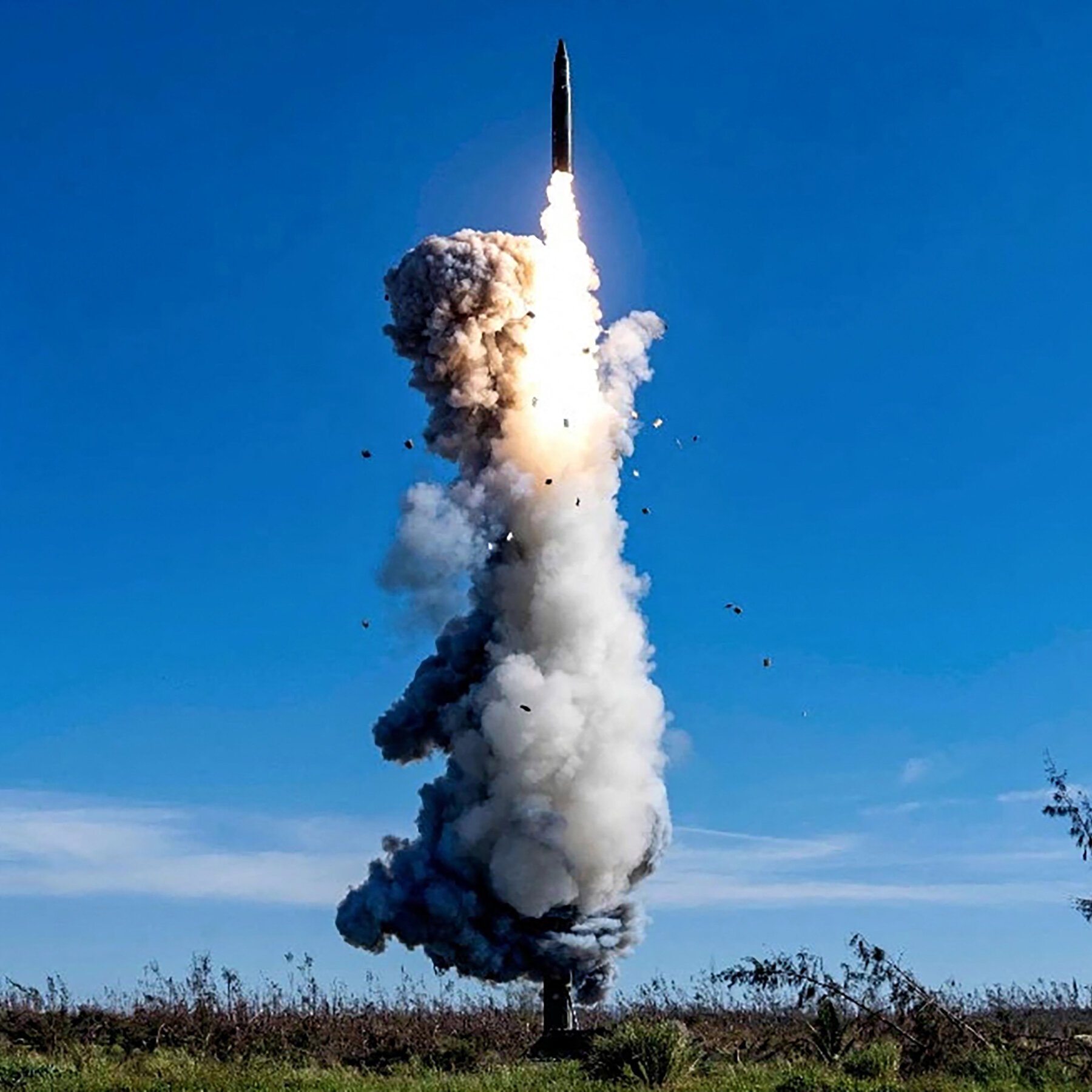 A missile launches upward from a grassy field, trailing a large plume of white smoke and bright flame against a clear blue sky.