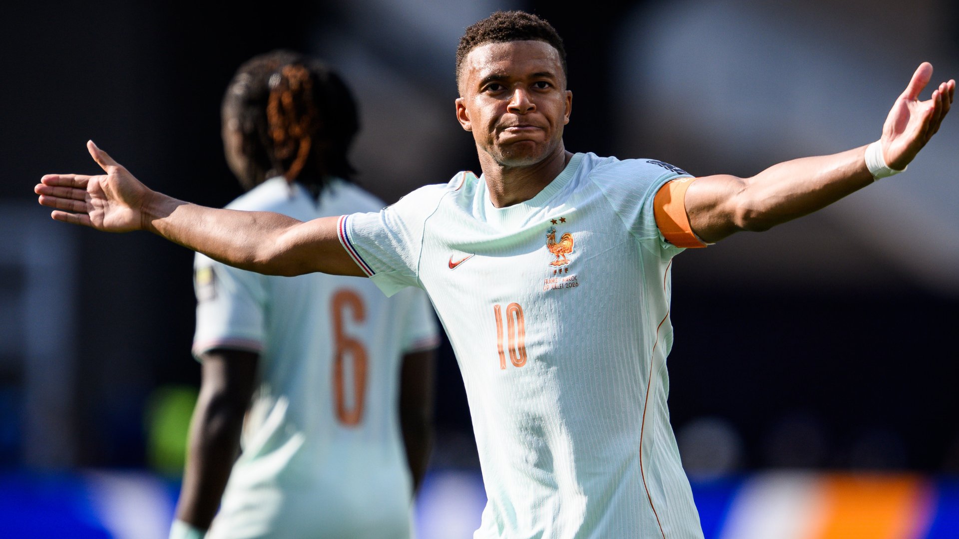 A soccer player in a white France national team jersey bearing the number 10 stands with both arms outstretched, wearing an orange captain's armband during a match.