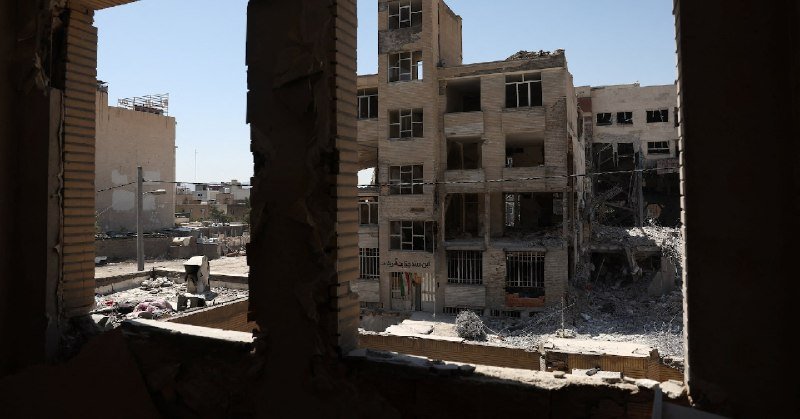 Viewed through a damaged building's interior, multiple heavily damaged multistory structures stand amid rubble-strewn ground under a clear sky.