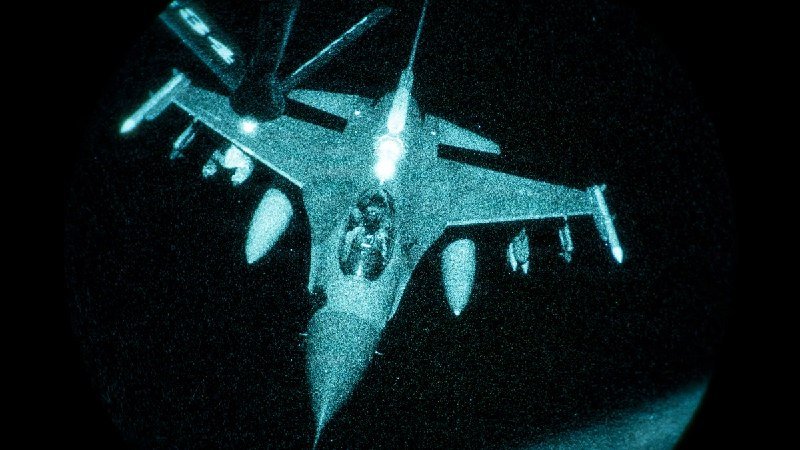 Night-vision image of a military fighter jet in flight, with a second aircraft visible in the upper left against a dark sky.