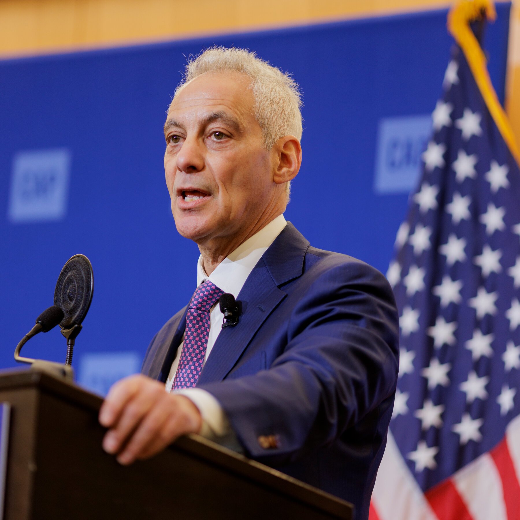 An older man with white hair speaks at a podium, wearing a navy suit and pink patterned tie, with microphones in front and an American flag visible.