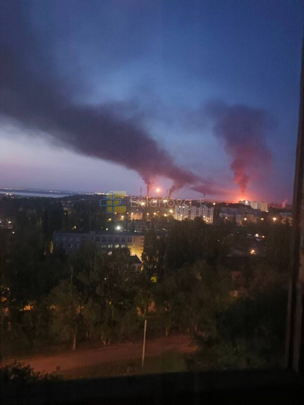 Thick smoke rises from illuminated buildings in a city at dusk, with trees and a river visible in the foreground.