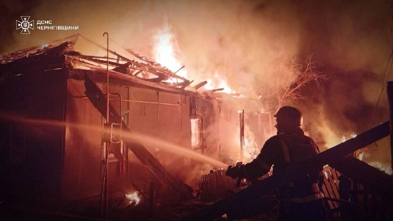 A firefighter in silhouette sprays water from a hose onto a large structure engulfed in flames and thick smoke, with an emblem and Cyrillic text visible in the corner.