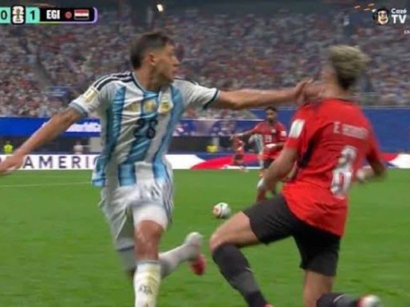 An Argentina player in a light blue and white striped jersey reaches toward a red-jerseyed player as they both move toward a soccer ball on a stadium pitch.