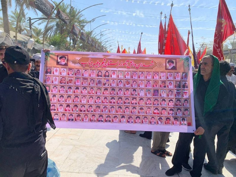 Two men hold a large banner displaying rows of portraits with Arabic script, while red flags and a security guard in black are visible in the background.