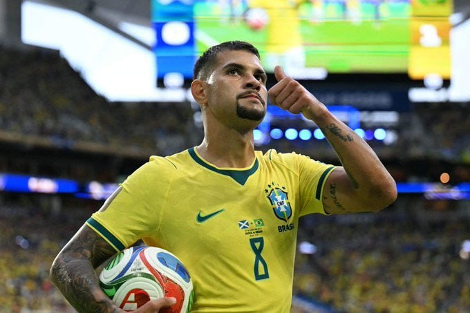 A soccer player in a yellow Brazil jersey (#8) holds a match ball under his arm and points upward with his index finger in a stadium.