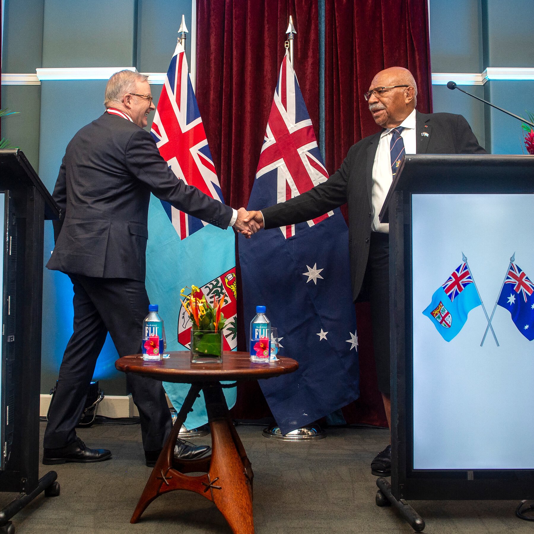 Two men in suits shake hands on a stage flanked by the Fiji and Australian flags, with a small table holding Fiji water bottles and flowers between them.