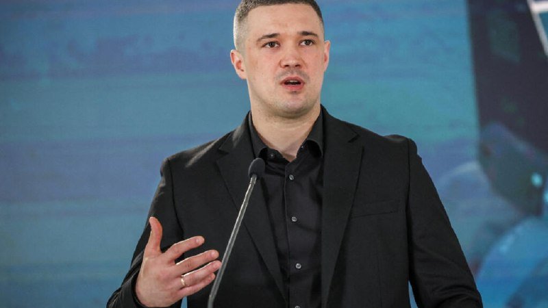 A man in a black suit and black shirt speaks at a microphone against a blue background, gesturing with his right hand.