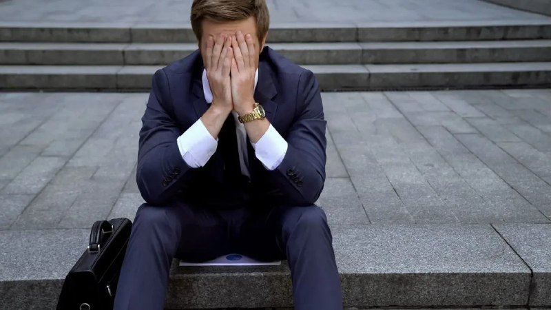 A man in a dark suit sits on outdoor stone steps with his face buried in both hands, a black briefcase beside him and a folder on his lap.