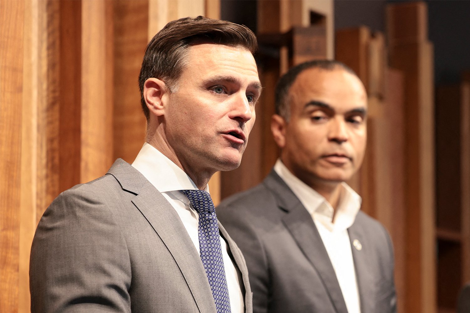 Two men in suits stand together against a wooden-paneled background, with the man in front appearing to speak.