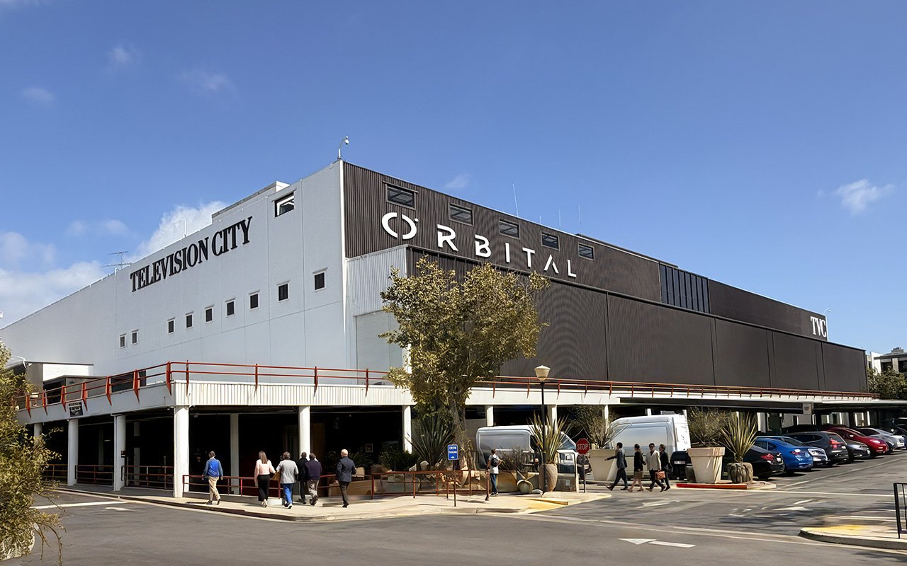 A large studio building labeled "TELEVISION CITY" and "ÖRBITAL" stands under a clear blue sky, with pedestrians walking nearby and cars parked along the side.