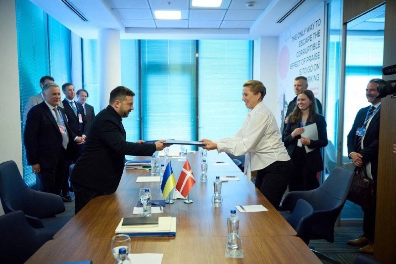 Two officials exchange a blue binder across a conference table displaying Ukrainian and Danish flags, with onlookers standing by in a modern meeting room featuring a wall-mounted informational graphic.