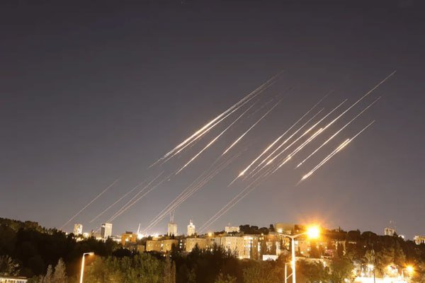 Bright streaks of light trail downward across the night sky above a lit city skyline.